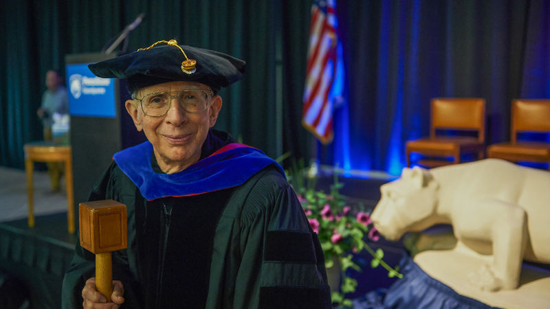 A man wears academic regalia and holds a mace.