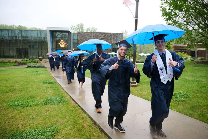 A group of students in commencement robes walks in the rain outside.