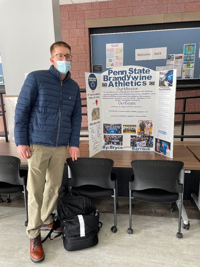 Bryce Barrouk wears a blue coat and stands in front of his poster detailing his internship with the Penn State Brandywine Athletics Department. 