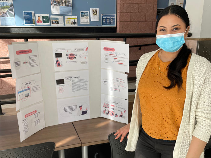 A student with dark hair and a goldenrod sweater stands in front of her poster at a recent event. 