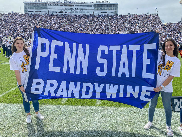 Two female students hold a Penn State Brandywine banner on the football field of Beaver Stadium.