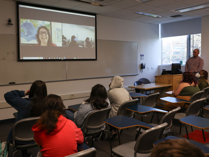 A woman is on a screen in a classroom speaking to a group of students.
