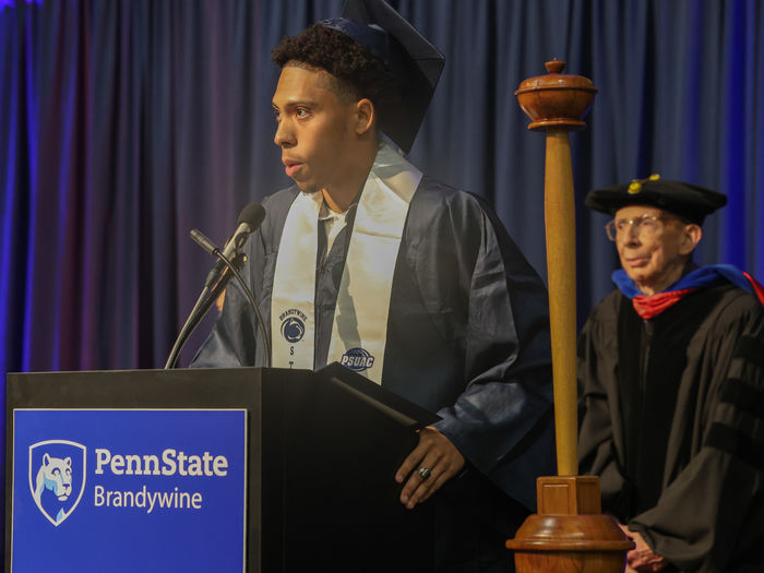 A male student wearing a cap and gown stands at a lectern speaking.