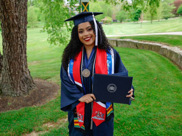 A person wearing a graduation gown stands outside holding her diploma.