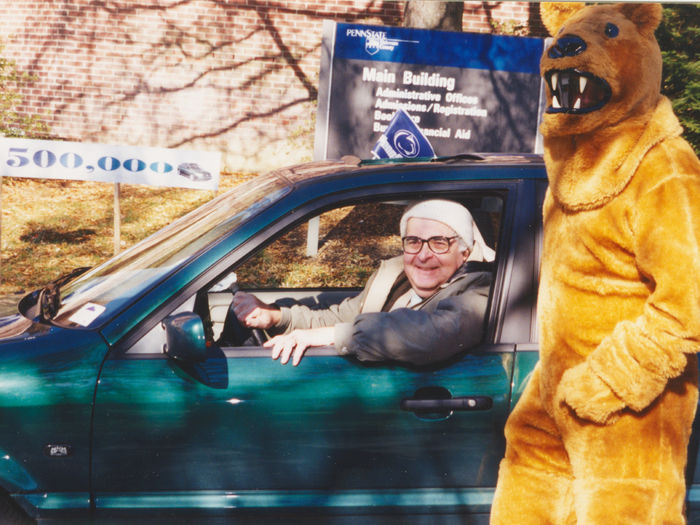 A man sits in a car with a lion mascot standing next to the car.