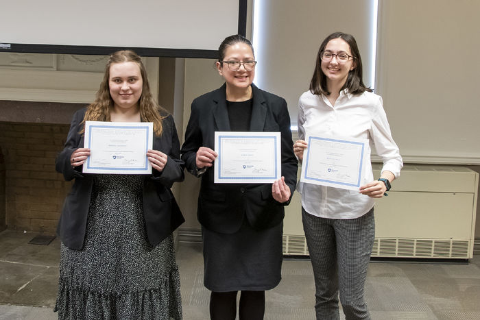 Three students stand in front of a fire place with certificates