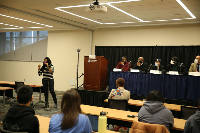 Panelists sit on the stage at a long table while a moderator facilitates a conversation with students, faculty and staff.  