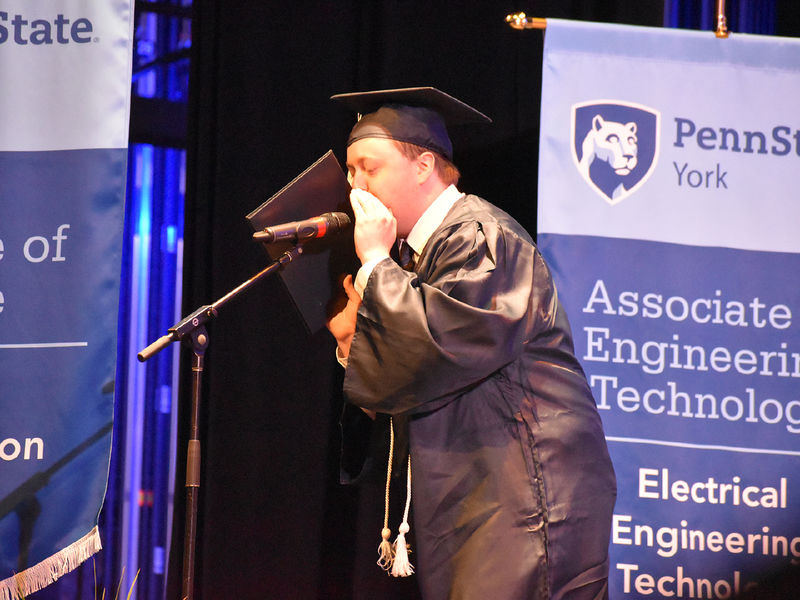 Male student in graduation cap and gown speaking into a microphone on a stabnd