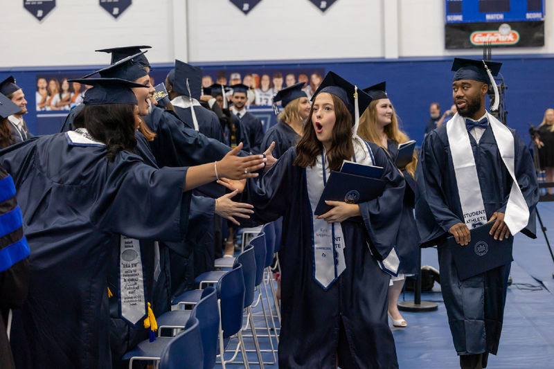 A graduate greets 2 friends as she exits after commencement. All are dressed in caps and gowns.