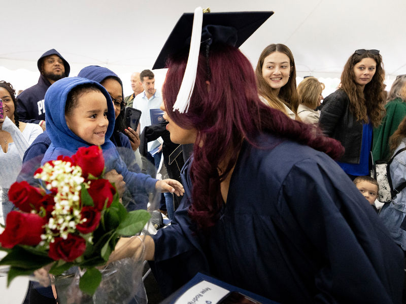 Woman in commencement gown bends down to accept a bouquet of flowers from a young boy.