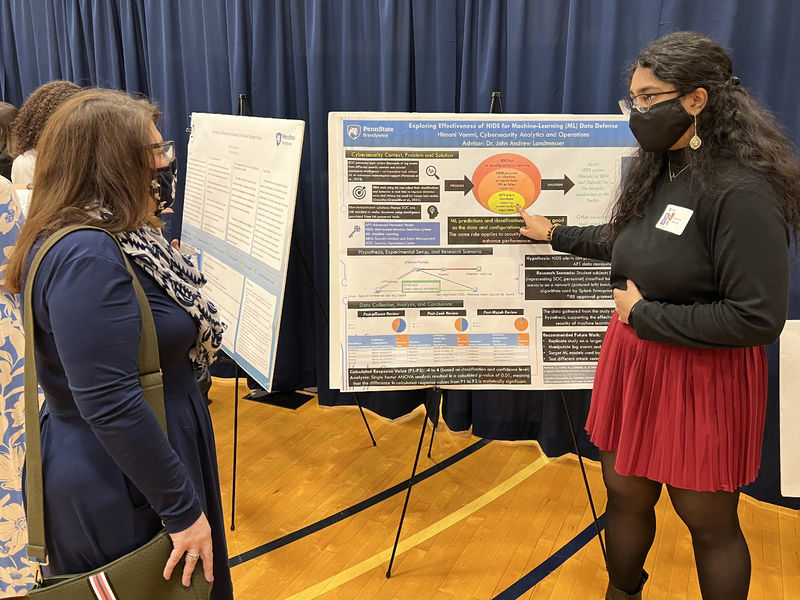 A women stands next to her research poster while explaining it to another woman.