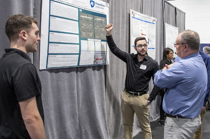 Student wearing black shirt points to research poster as he speaks with a symposium judge.