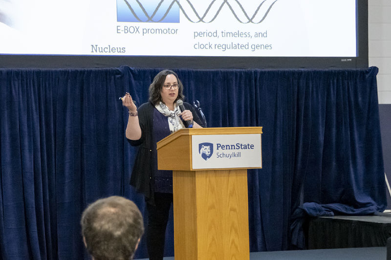 Woman stands in front of a projector screen and behind a podium, holding a microphone, as she gestures to her PowerPoint slide