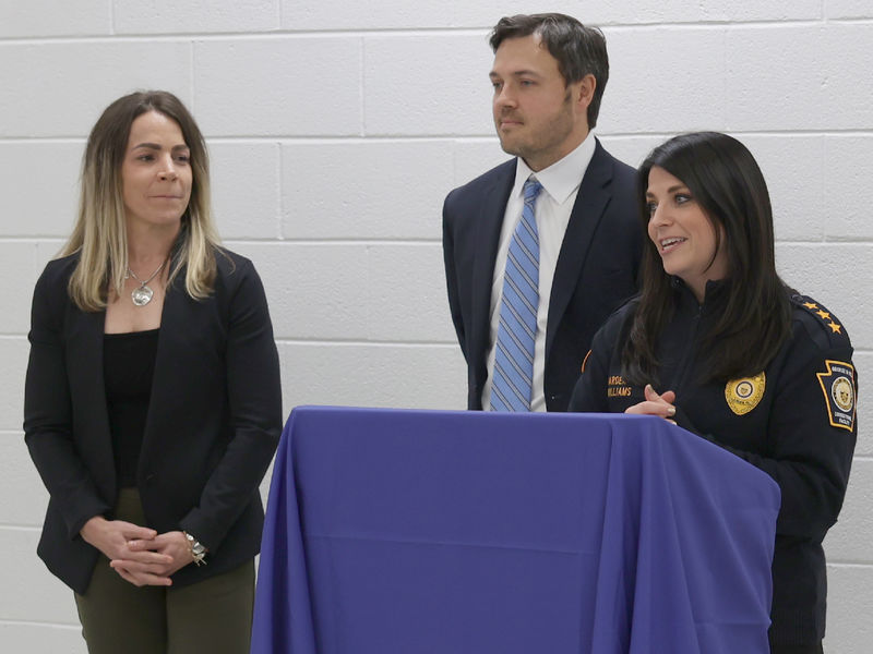 A woman speaks at a lectern with a man and another woman standing beside her.