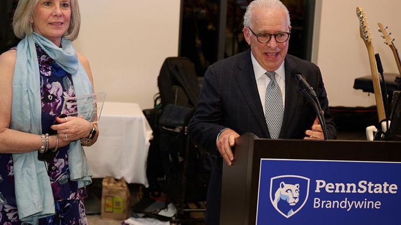 A man speaks at a lectern while a woman stands to his side holding an award.