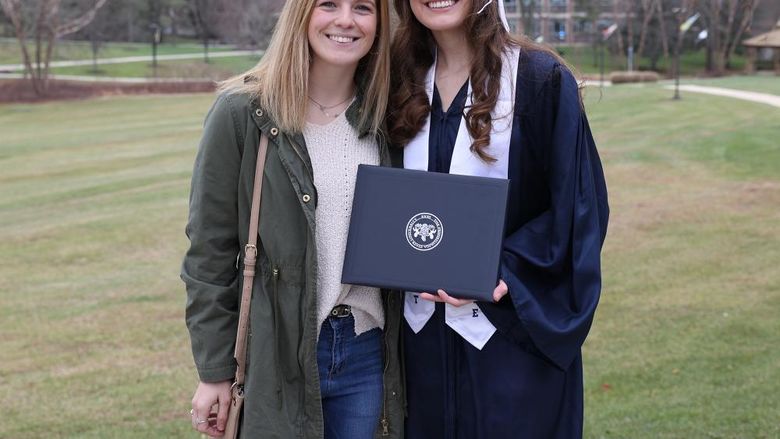 Two women stand outdoors. One is wearing a graduation cap and gown and holding a diploma cover.