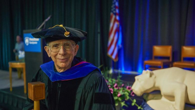 A man wears academic regalia and holds a mace.