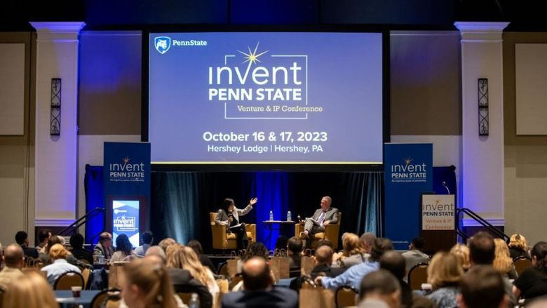 Dr. Karen Kim and Andrew Read speak sitting on a stage in front of an Invent Penn State Venture & IP Conference branded screen with a full audience in the foreground