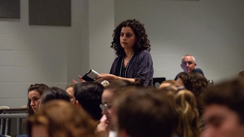 A woman stands to speak, surrounded by other students in an auditorium. 