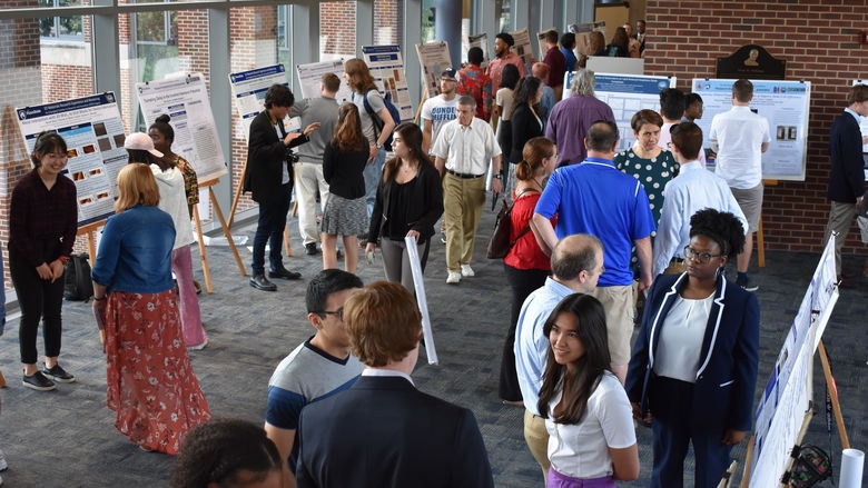 Scientific posters on easels line a large room, with windows on one side. People are grouped together in two or threes, discussing the research on the posters. 