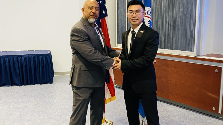 Two men shake hands in front of the flag of the United States of America and the Department of Homeland security. 
