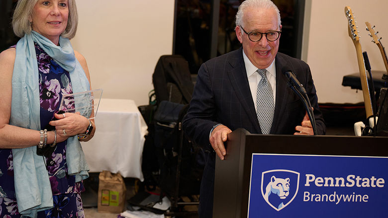 A man speaks at a lectern while a woman stands to his side holding an award.