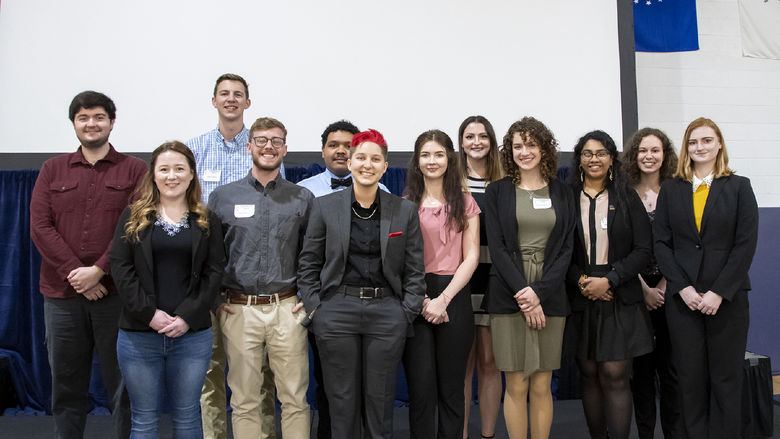 A dozen Penn State students pose for a photo on stage in front of a projector screen