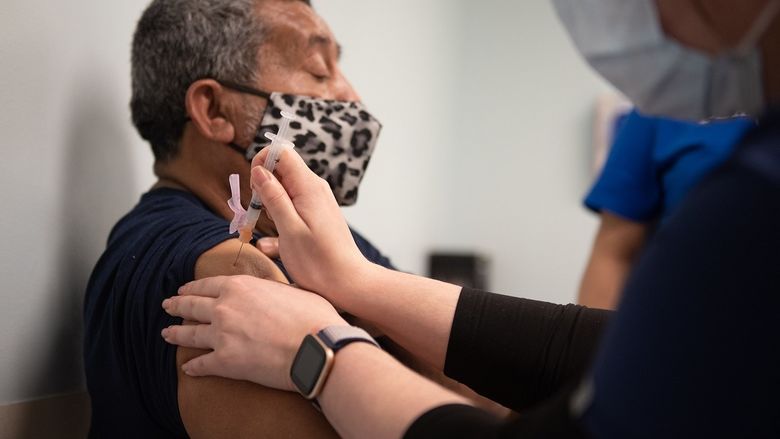 Samuel Garcia, who is seated and wears a mask, looks straight ahead as he receives a COVID-19 vaccine into his upper arm from a health care worker.