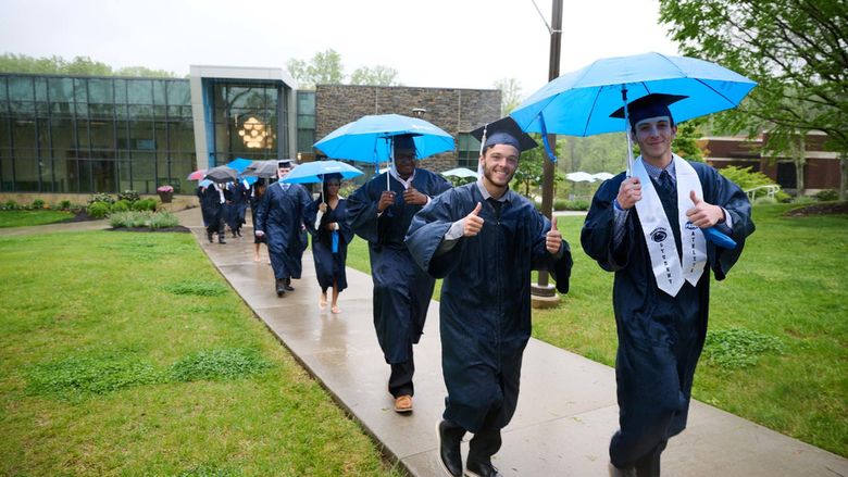 A group of students in commencement robes walks in the rain outside.