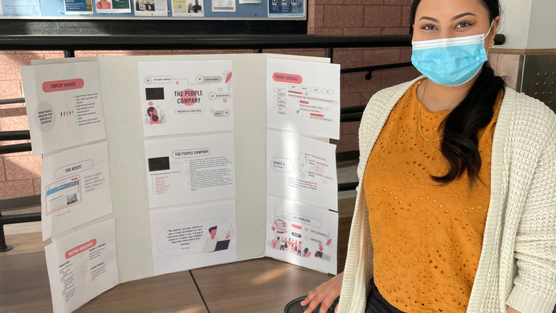 A student with dark hair and a goldenrod sweater stands in front of her poster at a recent event. 