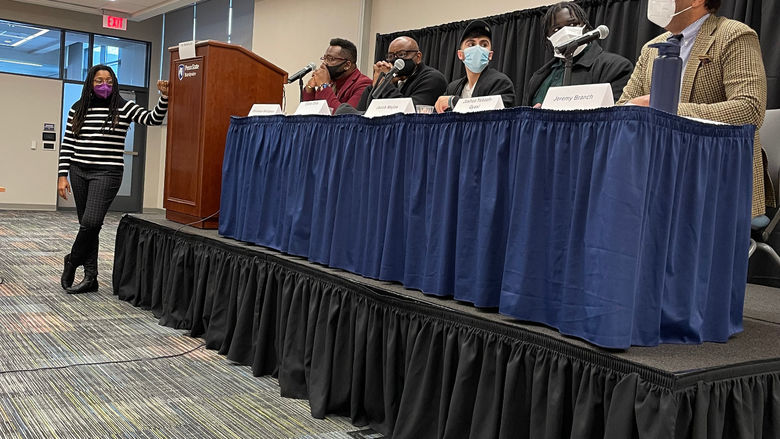 A group of men sit on a stage at a table with a blue table cloth with a woman standing beside the stage, moderating the discussion. 