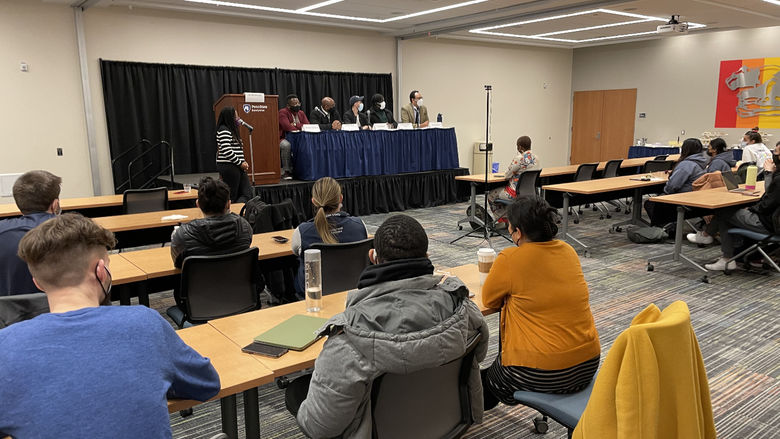 Community members seated at long wooden tables watch a group of panelists sitting on a stage with a facilitator standing in the foreground. 