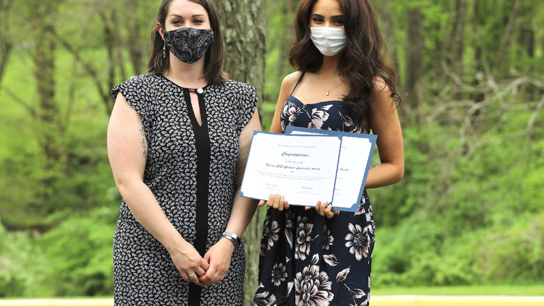 Two women stand outside with one of the women holding award certificates.