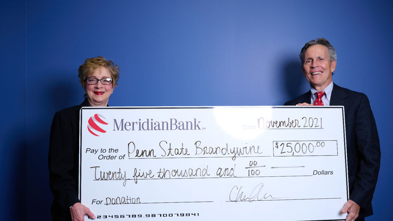 Meridian CEO Chris Annas with Penn State Brandywine Chancellor Marilyn Wells stand in front of a blue wall holding a large white check. 