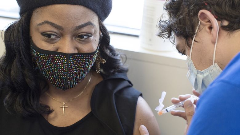 Tamika Washington, who wears a jeweled face mask, a cross necklace and a beret, looks away from her bare arm as a health care worker prepares to insert a syringe filled with the COVID-19 vaccine.