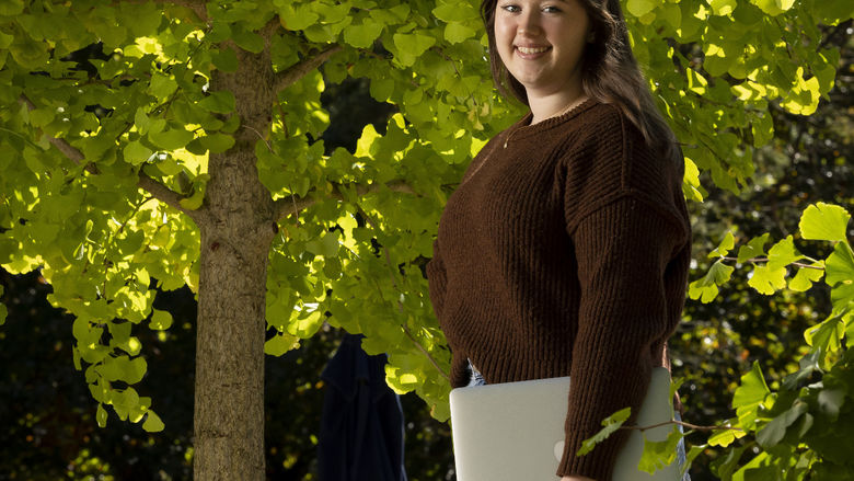 A female student stands outside holding a laptop.