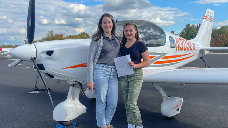 Two women stand next to a small plane holding a certificate.