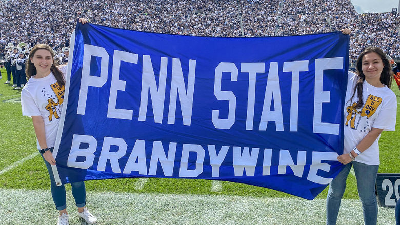 Two female students hold a Penn State Brandywine banner on the football field of Beaver Stadium.