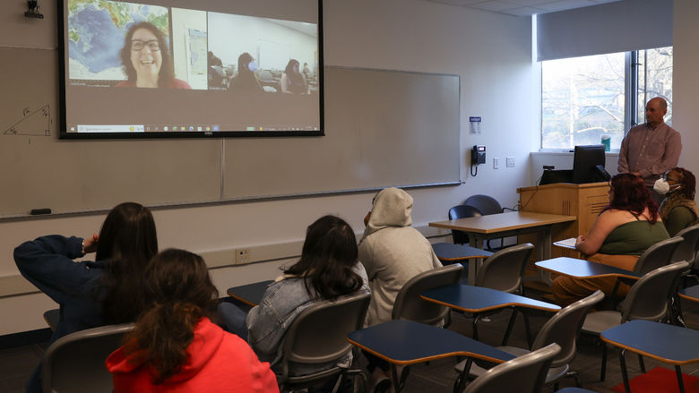 A woman is on a screen in a classroom speaking to a group of students.