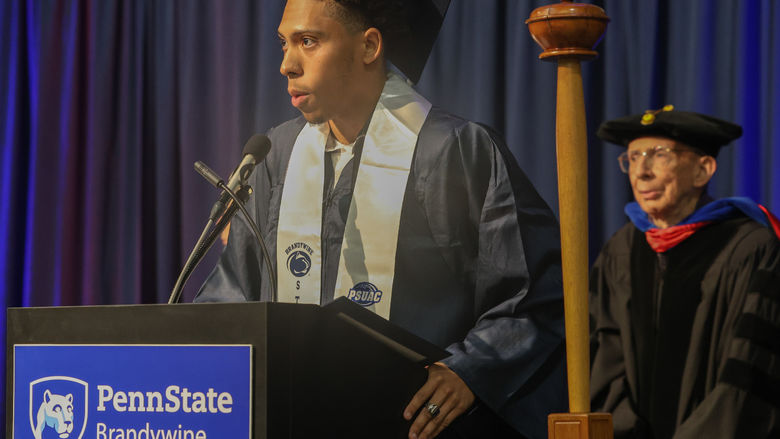 A male student wearing a cap and gown stands at a lectern speaking.