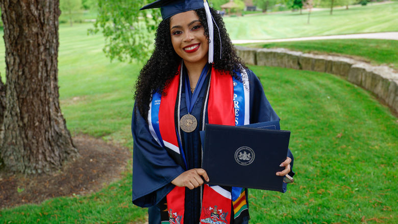 A person wearing a graduation gown stands outside holding her diploma.