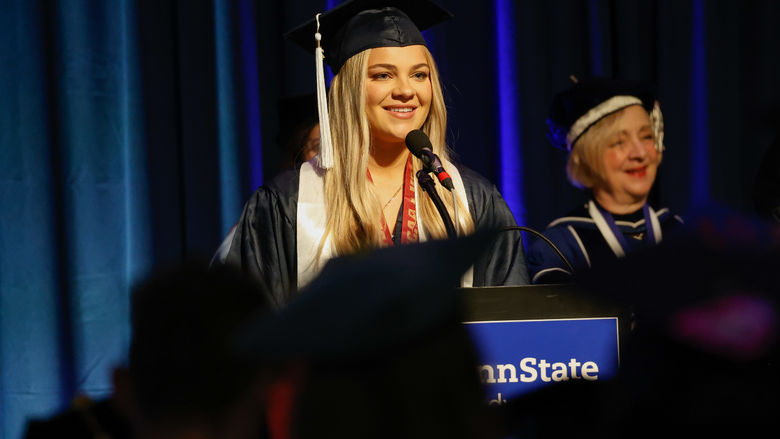 A person wearing a graduation gown stands at a lectern to speak at commencement.