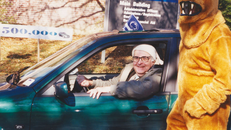 A man sits in a car with a lion mascot standing next to the car.
