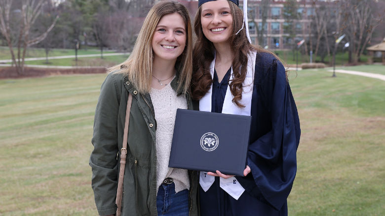 Two women stand outdoors. One is wearing a graduation cap and gown and holding a diploma cover.
