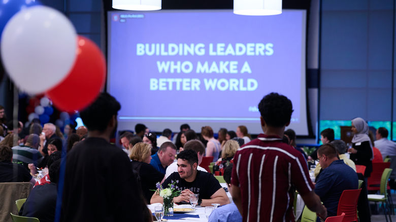 crowd sitting at tables with "building leaders who make a better world" on projector