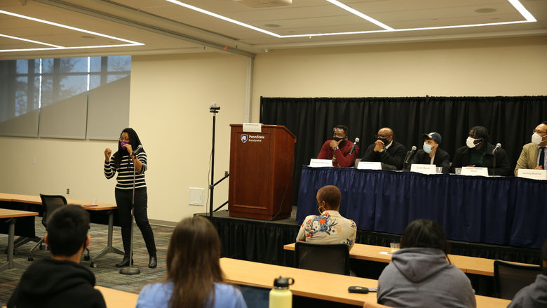 Panelists sit on the stage at a long table while a moderator facilitates a conversation with students, faculty and staff.  