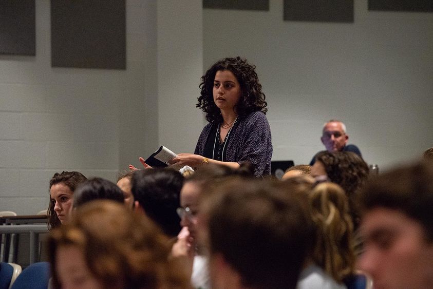A woman stands to speak, surrounded by other students in an auditorium.