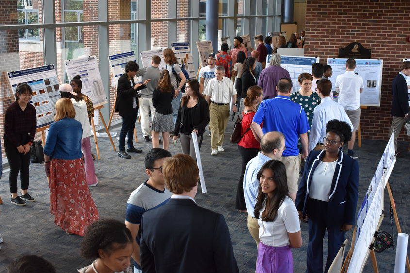 Scientific posters on easels line a large room, with windows on one side. People are grouped together in two or threes, discussing the research on the posters.