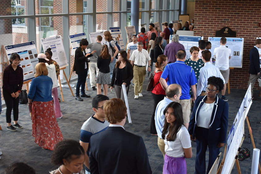 Scientific posters on easels line a large room, with windows on one side. People are grouped together in two or threes, discussing the research on the posters. 