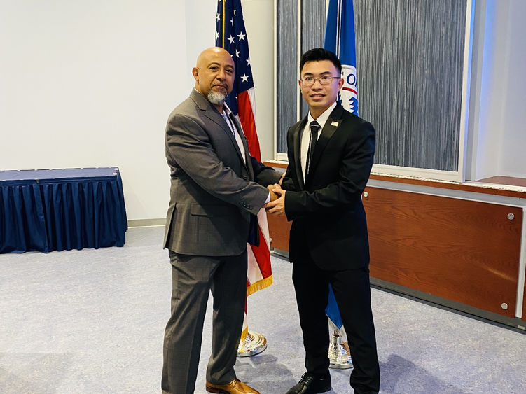 Two men shake hands in front of the flag of the United States of America and the Department of Homeland security. 